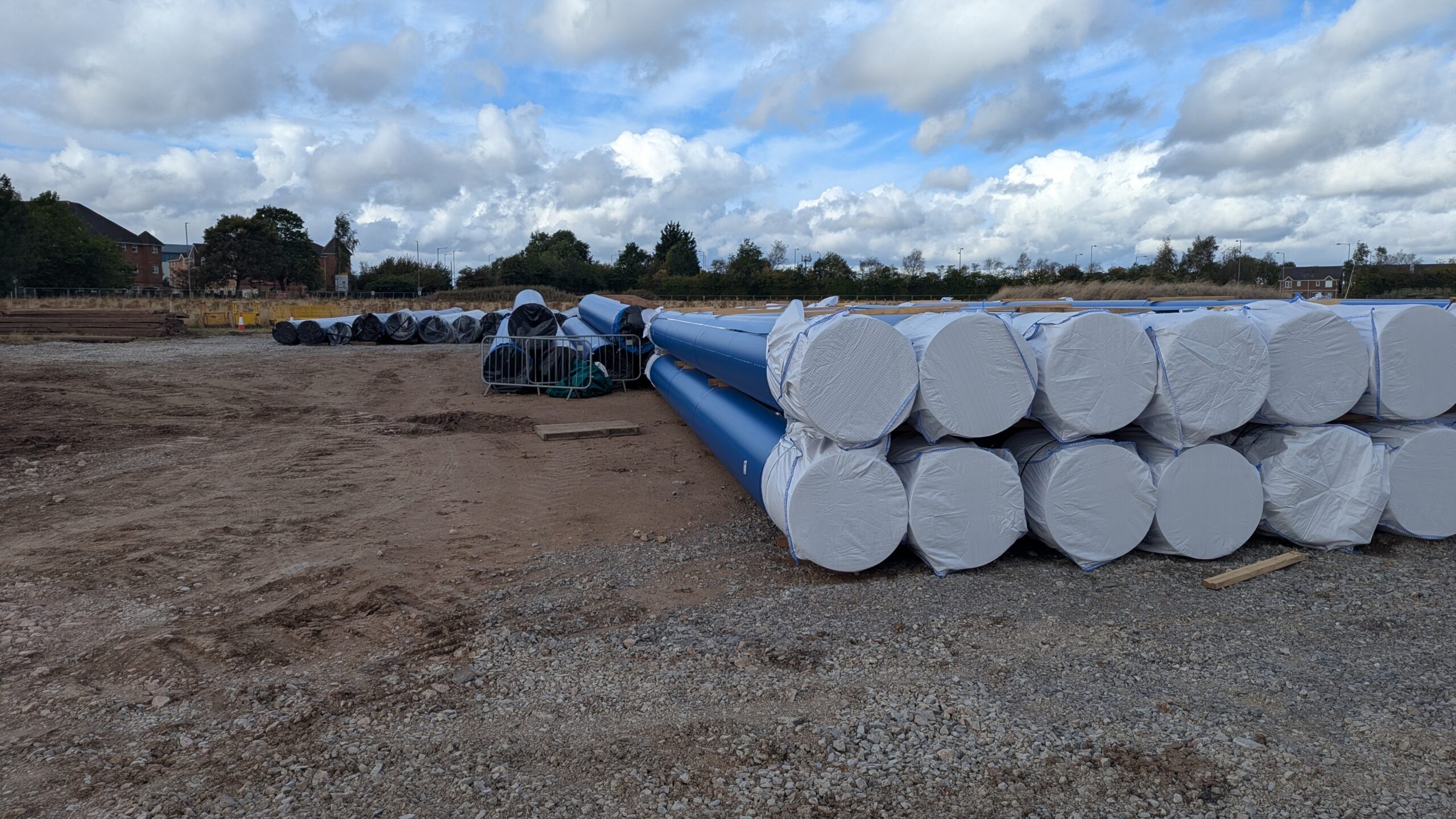 A pipe yard on site: wrapped pipe bundles and lengths of blue PE pipe under a wide sky