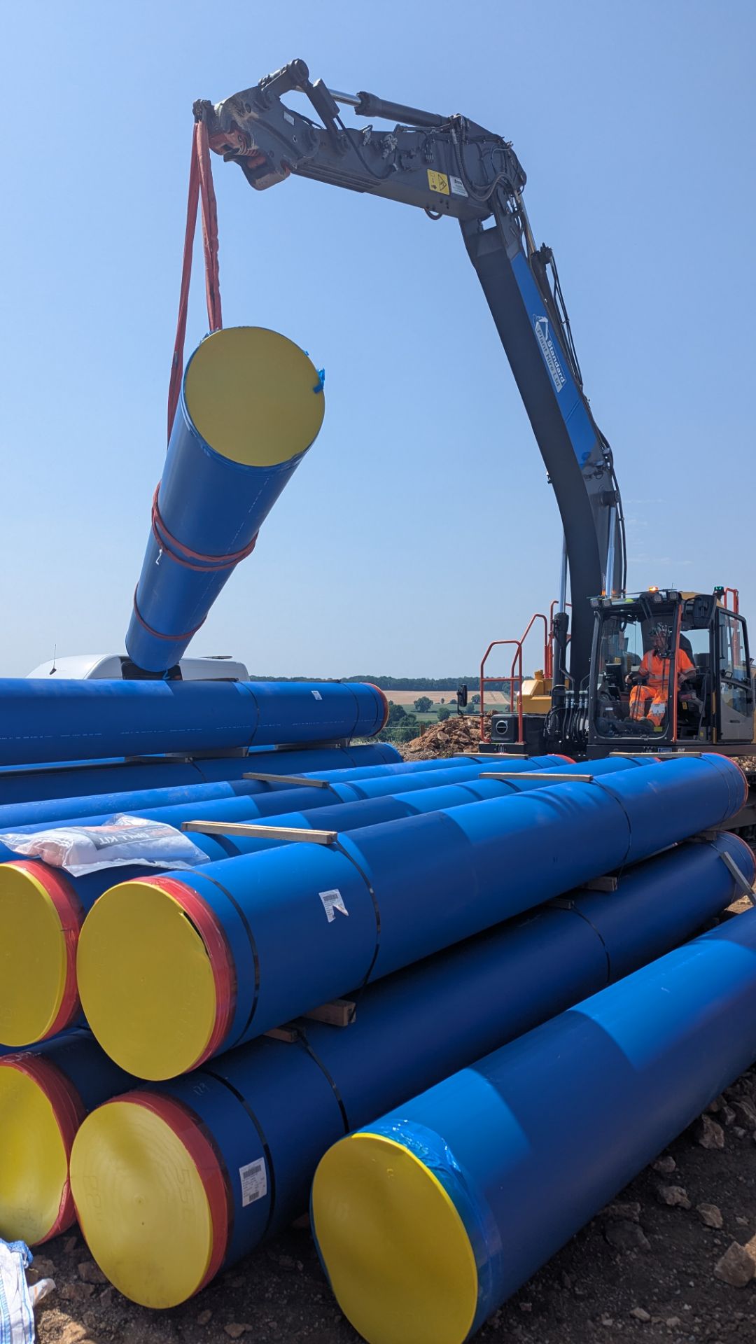 An excavator lifting a length of blue PE pipe off a stack on site